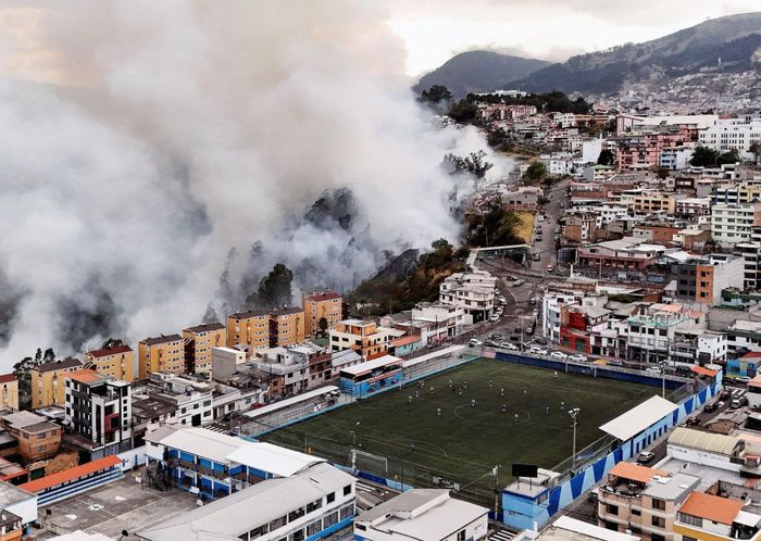 Một trận cháy rừng bùng lên trên sườn đồi ở Quito, Ecuador, ngày 13/9, khói cuồn cuộn phía sau khu dân cư trong lúc một nhóm người vẫn chơi bóng đá gần đó.