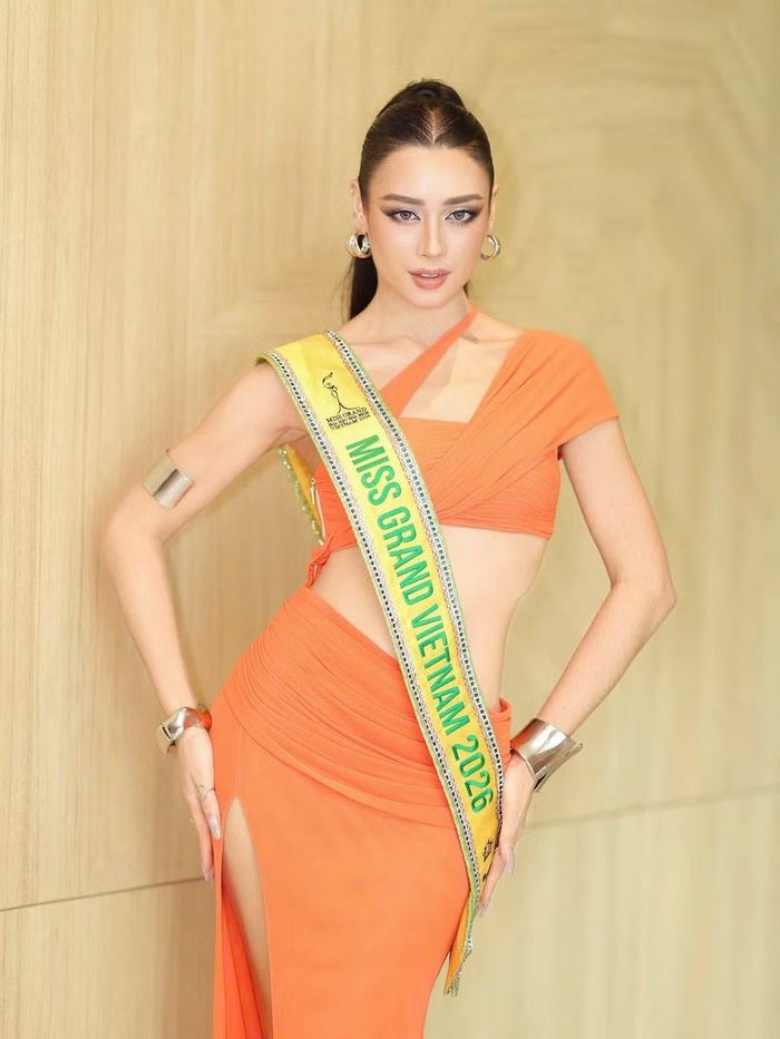 Emoura Phạm diện trang phục màu cam với thiết kế bất đối xứng, khoảng hở ở eo khi tham gia hoạt động tại Miss Grand International 2026. Cô kết hợp tóc đuôi ngựa buộc cao, khuyên tai kim loại và lối trang điểm sắc nét.