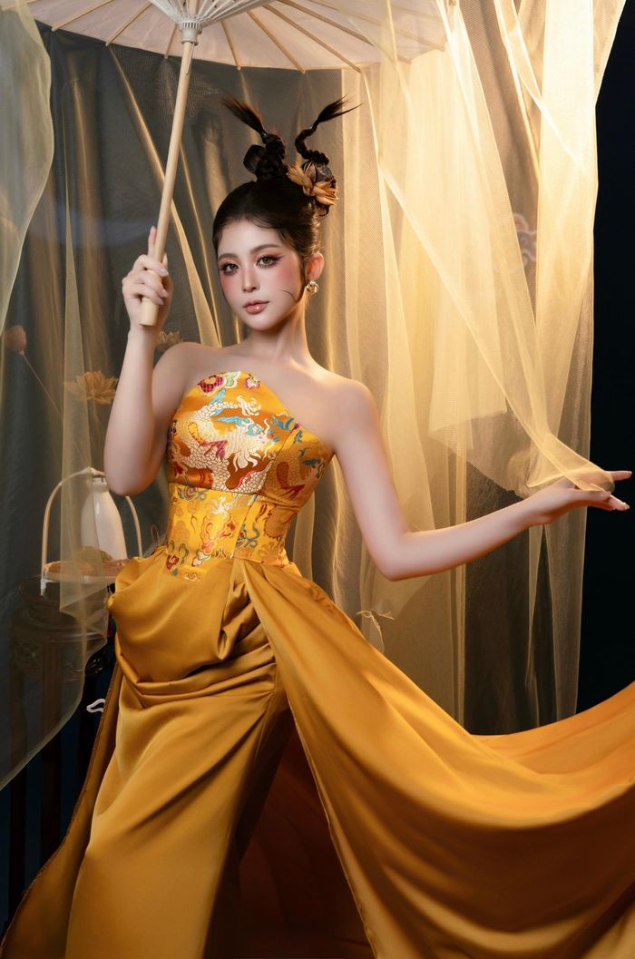 Ở loạt ảnh sau, Thùy Trang khoác lên mình thiết kế màu vàng đồng với họa tiết rồng, kết hợp kiểu tóc búi cao và phụ kiện lấy cảm hứng từ hoa sen, hạt sen. Sắc vàng nổi bật trên nền xanh đậm cùng ánh sáng ấm khiến tổng thể bộ ảnh trở nên rực rỡ hơn. Hình ảnh chiếc bánh Trung thu được đưa vào một số khung hình như điểm nhấn gợi nhắc về mùa đoàn viên.