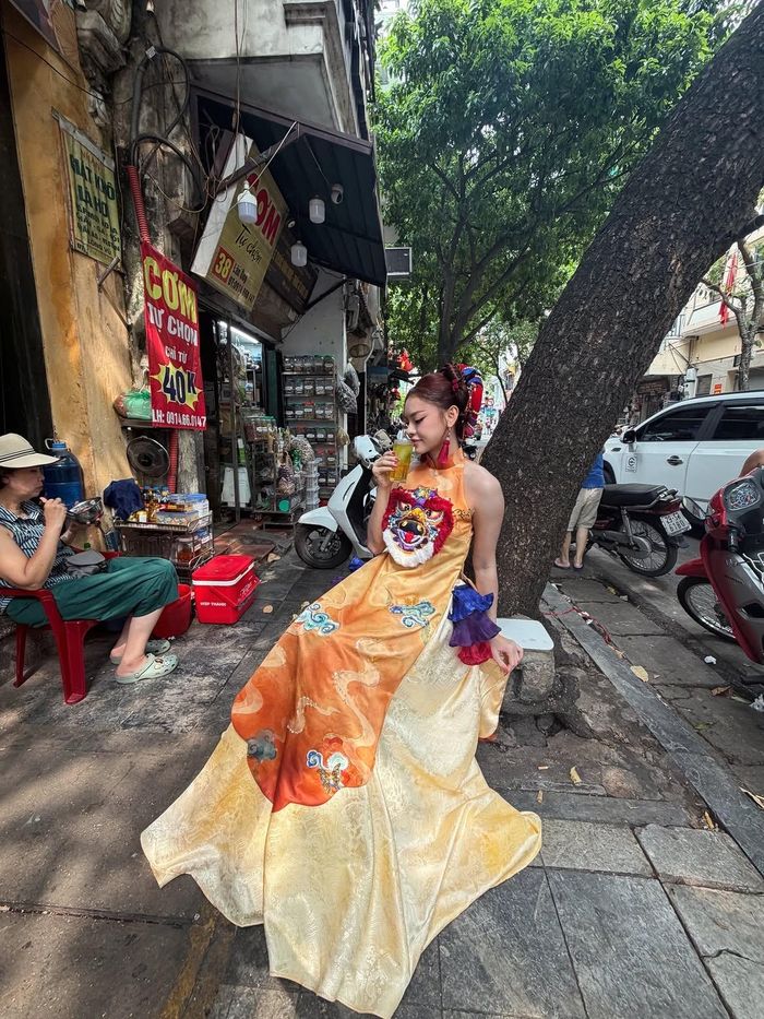 Nếu Thanh Thanh Huyền và Bạch Lưu Dương khai thác layering theo hướng giữ nguyên cấu trúc áo dài, người mẫu Mai Ngô lại lựa chọn một thiết kế có nhiều chi tiết cách tân hơn. Người mẫu diện áo dài cổ cao, không tay, màu cam nổi bật khi chụp hình Trung thu trên đường phố Hà Nội. Phần tà áo được trang trí bằng họa tiết mây trời và hình đầu lân. Bên dưới là phần chân váy bằng gấm màu vàng mỡ gà, điểm hoa văn chìm. Việc kết hợp nhiều chất liệu và chi tiết trang trí giúp bộ trang phục giữ được hình dáng quen thuộc của áo dài nhưng có thêm nét lễ hội, phù hợp không khí Trung thu. Ảnh: @maingo.official.