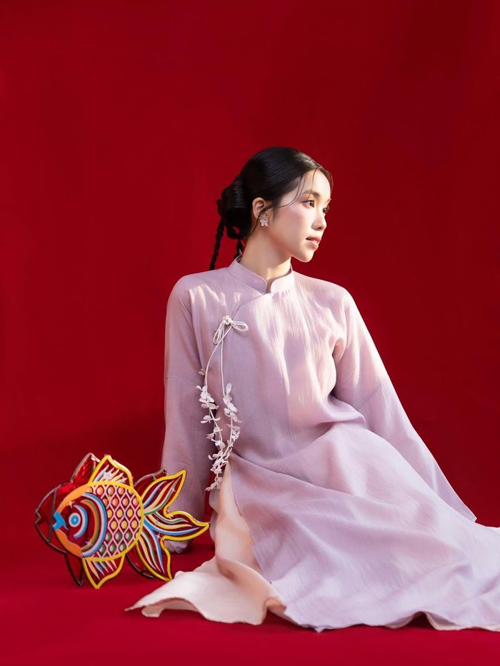 Trong khi đó, influencer Trịnh Hà Vi chọn cách tiết chế hơn với áo dài dáng suông, cổ cao, tông tím pastel. Màu sắc nhẹ cùng phom dáng đơn giản tạo hình ảnh thanh lịch, dịu dàng và nền nã. Điểm nhấn nằm ở dải hoa 3D được xử lý như những cánh hoa rơi, tạo hiệu ứng bay bổng trên thân áo. Hà Vi kết hợp thiết kế với kiểu tóc búi cầu kỳ và những món đồ chơi quen thuộc trong dịp Trung thu làm đạo cụ. Cách lựa chọn này đưa không khí lễ hội vào bộ ảnh mà không cần sử dụng quá nhiều chi tiết trên trang phục. Ảnh: @_trinhhavi_.