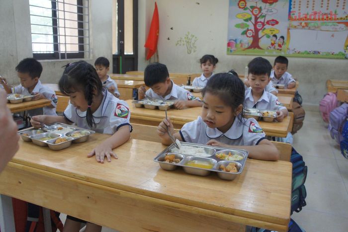Tại lớp 1, giáo viên chủ nhiệm tận tình hướng dẫn các em cách cầm thìa, chia thêm cơm canh cho những em ăn khỏe và động viên các bé biếng ăn hoàn thành bữa trưa.