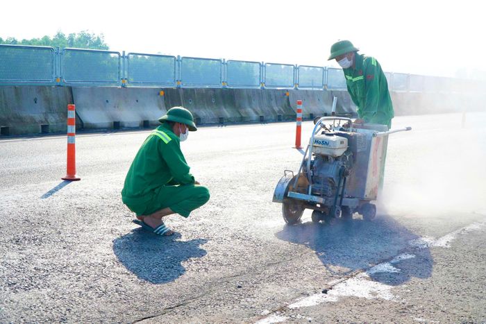 Một nhóm công nhân trực tiếp xử lý mặt đường, nhóm khác rà soát phạm vi cần bóc dỡ, trong khi lực lượng phía ngoài tập trung thực hiện các công việc phục vụ thi công và bảo đảm an toàn giao thông. Từng phần mặt đường hư hỏng được bóc bỏ, để lộ kết cấu bên dưới phục vụ kiểm tra, đánh giá trước khi thực hiện các bước xử lý tiếp theo.