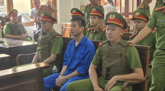 Bị cáo mặc áo xanh tại phiên tòa, bên cạnh các cán bộ làm nhiệm vụ bảo vệ.
