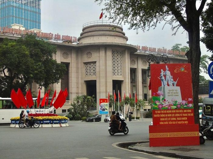 Trước trụ sở Ngân hàng Nhà nước Việt Nam, những lá cờ đỏ được bố trí dọc khu vực phía trước công trình, nổi bật bên cạnh cụm hoa trang trí và bảng tuyên truyền.