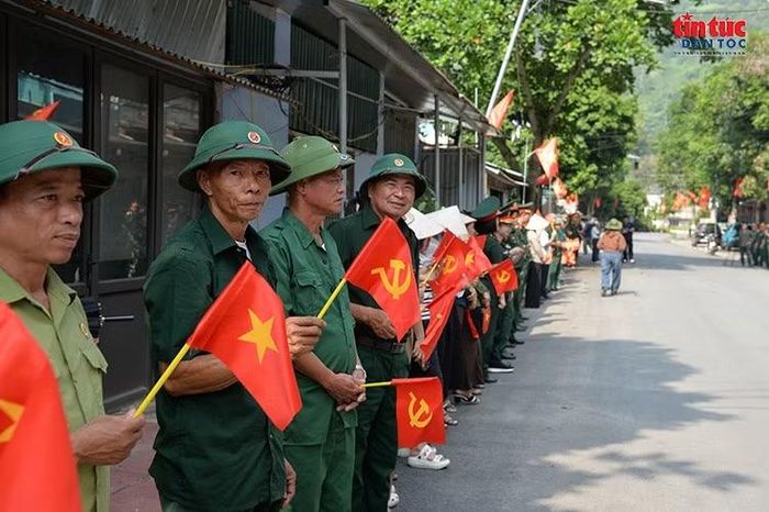 Các cựu chiến binh trong trang phục màu xanh, cầm cờ Tổ quốc và cờ Đảng, đứng chờ đoàn xe chở hài cốt liệt sĩ đi qua, tiếp nối nghĩa tình đồng đội nơi biên cương.