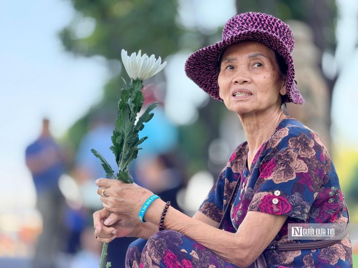 Một cụ bà ngồi bên vệ đường, tay cầm nhành hoa trắng chờ đợi đoàn xe chở các anh hùng dân tộc đi qua. (Ảnh: Hoàng Hùng)