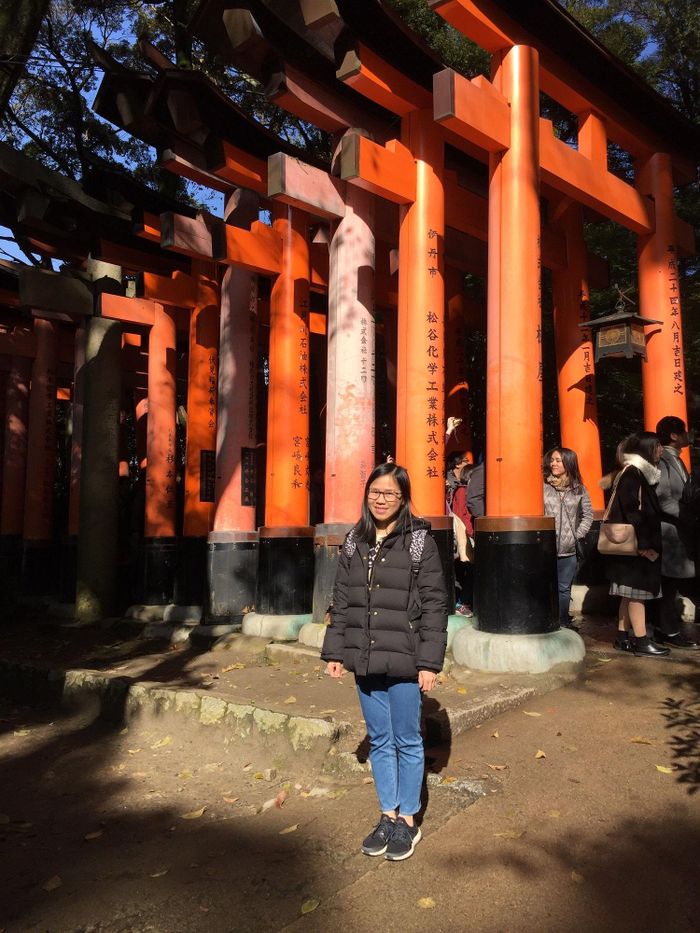 Chị Tiếng tại Đền Fushimi Inari Taisha (Kyoto). Phía sau là các cổng Torii màu cam (Senbon Torii) đặc trưng với các dòng chữ Hán (Kanji) khắc tên người/doanh nghiệp công đức.