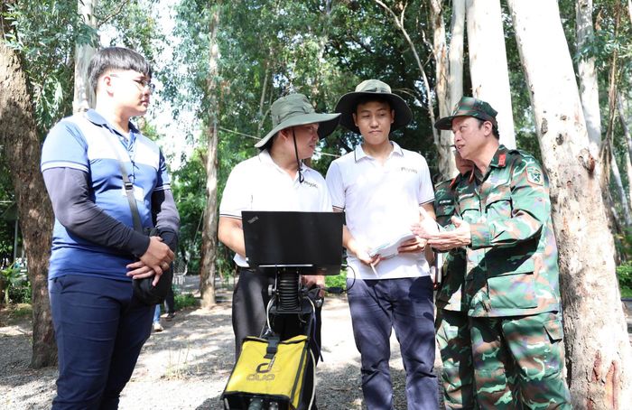  Đại tá Nguyễn Đình Chuẩn (Phó Chính Ủy Bộ Tư lệnh TPHCM, bìa phải) trao đổi công việc khảo sát dò tìm mộ Liệt sĩ với nhóm đo đạc ra đa xuyên đất (Bộ môn Vật lý địa cầu, Khoa Vật lý – Vật lý kỹ thuật, Trường ĐH Khoa học tự nhiên, ĐHQG TPHCM) ở công viên Lê Thị Riêng, tháng 7/2026. 