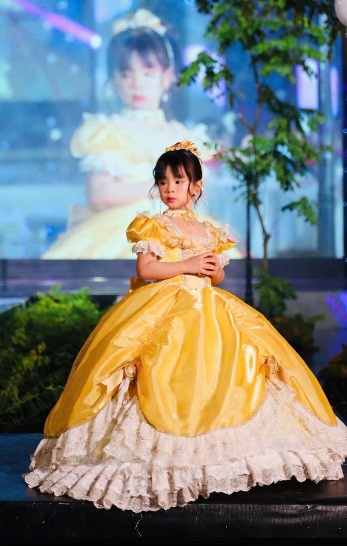 Mẫu nhí trong bộ váy công chúa vàng rực rỡ, kết hợp chân váy ren trắng và vương miện nhỏ, duyên dáng tạo dáng trên sàn diễn với vẻ hồn nhiên, đáng yêu.