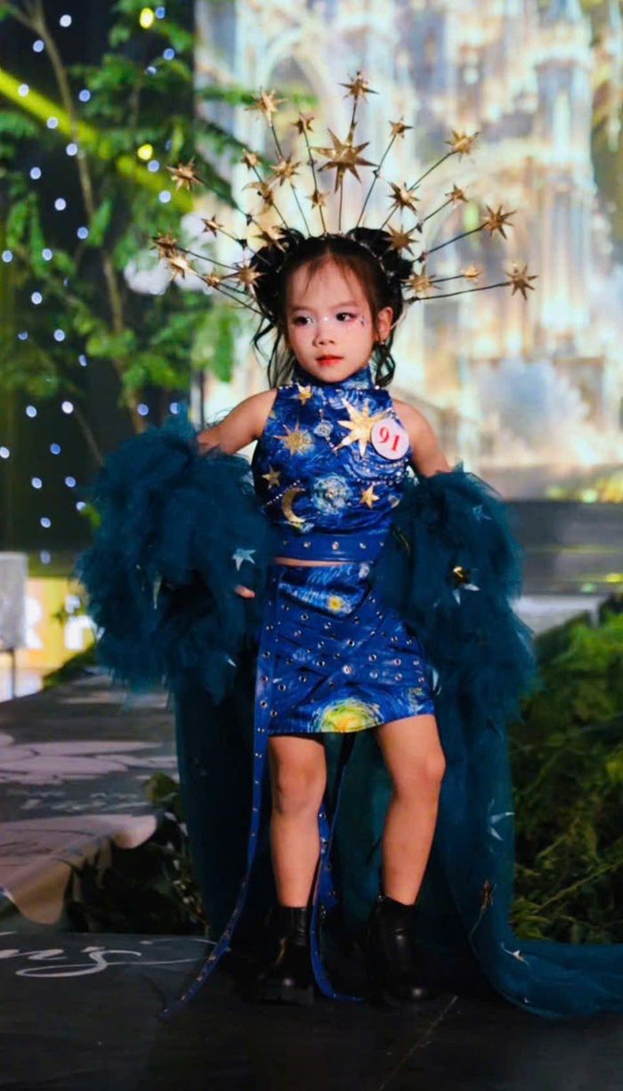 Mẫu nhí số báo danh 91 nổi bật với thiết kế thời trang lấy cảm hứng từ vũ trụ, kết hợp sắc xanh huyền bí, họa tiết các vì sao và áo choàng lông vũ xanh đậm. Chiếc vương miện ngôi sao độc đáo tạo điểm nhấn ấn tượng, mang đến hình ảnh vừa cá tính, vừa huyền ảo trên sàn diễn Vietkids Junior Fashion Week.