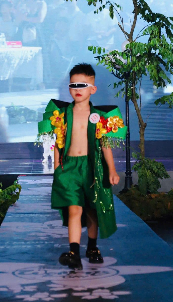 Bộ trang phục xanh lá nổi bật với thiết kế cầu vai góc cạnh, họa tiết hoa rực rỡ và những dải tua rua lấp lánh. Kết hợp kính mắt hiện đại, mẫu nhí mang đến phong cách thời trang phá cách, năng động và đầy cá tính trên sàn diễn.
