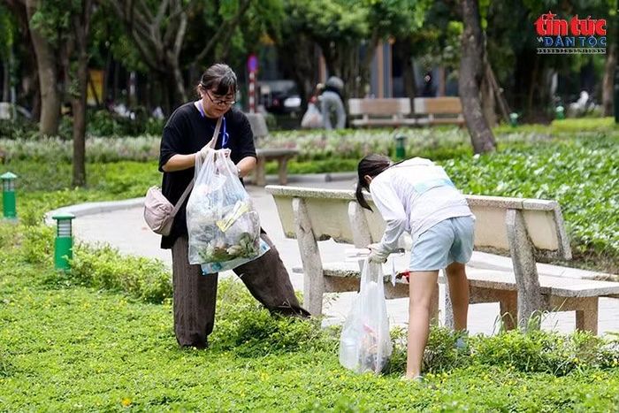 Các thành viên phối hợp thu gom rác quanh ghế đá và thảm cỏ trong khuôn viên xanh.