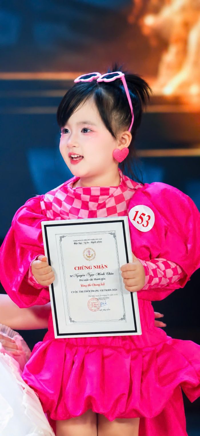 Mẫu nhí Nguyễn Ngọc Minh Châu rạng rỡ trong trang phục hồng nổi bật, cầm trên tay giấy chứng nhận “Vòng thi Chung kết” cuộc thi thời trang Vietkids năm 2024.