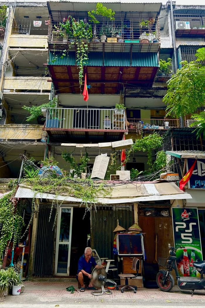 Dưới những tầng ban công phủ đầy cây cối và những lá cờ đỏ rực rỡ, người thợ già lặng lẽ, tỉ mỉ sửa chiếc quạt máy đã cũ