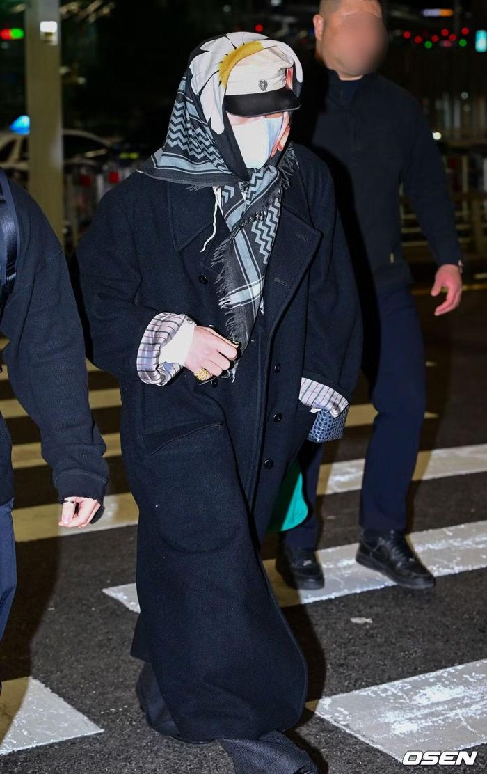 G-Dragon xuất hiện với phong cách thời trang sân bay đầy lịch lãm, diện chiếc áo khoác dài màu đen phối cùng quần jeans ống rộng. Cách quàng khăn đặc trưng của anh cũng là một điểm nhấn nổi bật.