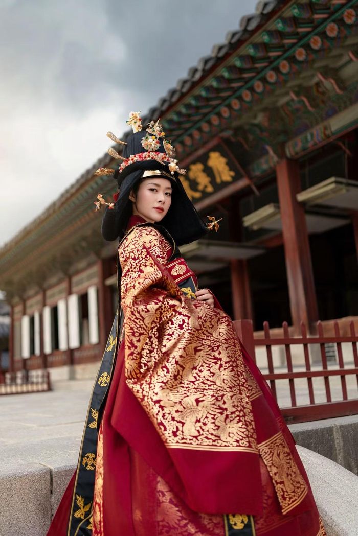Đứng trước cánh cửa cung điện, nữ diễn viên khoác lên mình bộ lễ phục đỏ thêu hoa văn vàng nổi bật. Dáng đứng ngay ngắn, hai tay đặt trước người cùng vẻ mặt nghiêm nghị của cô khiến khung hình gợi nhớ đến những cảnh trong phim cổ trang.