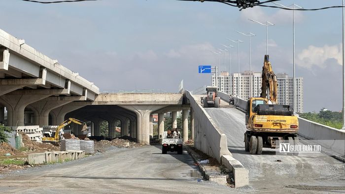 Công trường nhộn nhịp tiếng máy móc chuyên dụng hoạt động liên tục dọc theo các nhánh dầm cầu đang hoàn thiện lắp lan can, biển báo và dựng cột đèn chiếu sáng.