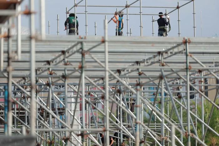 Tại khu vực giàn giáo, nhóm công nhân phía dưới phối hợp với những người làm việc trên cao. Các thanh ngang, cột đứng lần lượt được đưa vào vị trí. Hệ khung tiếp tục được nối cao bên cạnh phần mái che đã dựng.
