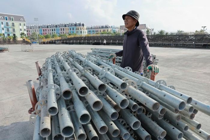 Các thanh giàn giáo được xếp dọc khu vực thi công. Công nhân vận chuyển đến vị trí lắp dựng. Phía sau là hệ khung đã được dựng cao, tạo nên những hình dáng đầu tiên của sân khấu chung kết.
